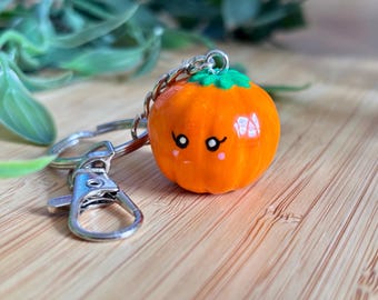 Può includere: Un portachiavi a forma di zucca arancione con un viso sorridente e un gambo verde. Il portachiavi ha un moschettone e una catena argentati. Su una superficie di legno con sfondo di fogliame verde.