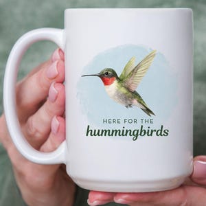 May include: White ceramic mug featuring a watercolor hummingbird design with the text "HERE FOR THE hummingbirds" in green. The hummingbird has red, green, and yellow details. The mug has a large handle.
