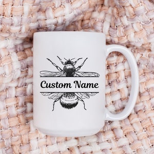 May include: White ceramic mug with a black bee illustration and the text "Custom Name". The mug has a large handle and is set against a textured, neutral-toned background. The bee design is split by a horizontal line for personalization.