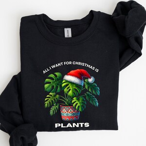 May include: Black sweatshirt with a graphic of a Monstera plant wearing a Santa hat. The text reads "ALL I WANT FOR CHRISTMAS IS PLANTS" in white. The plant is in a decorative pot with a red, blue, and white pattern.