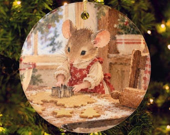 Op de afbeelding: Een ronde keramische kerstversiering met een bruine muis in een rood schort met witte sneeuwvlokpatronen, staand aan een houten tafel en een stervormige metalen koekjesvorm gebruikend op uitgerold deeg met bloem.