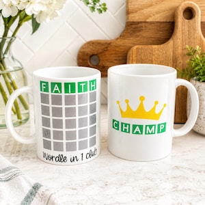 May include: Two white ceramic mugs. The left mug features a grid with the word FAITH in green boxes and the text Wordle in 1 club below. The right mug displays a yellow crown graphic above the word CHAMP in green boxes with white lettering.