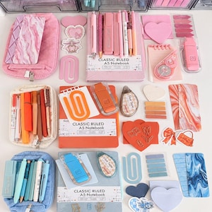 May include: A collection of stationery items organized by color, including three A5 notebooks in pink, orange, and blue, matching pencil cases, pens, highlighters, glue tape dispensers, heart-shaped sticky notes, and various paper clips.