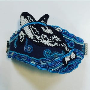 Op de afbeelding: Een kralenarmband met een zwart-witte orka die zwemt in blauwe en turquoise golven. De armband is gemaakt van kleine kralen en heeft een zilveren sluiting.