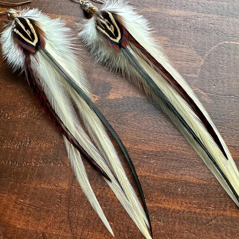 May include: A pair of long feather earrings with white, black, and red feathers. The earrings are made with natural feathers and have a simple gold wire design.