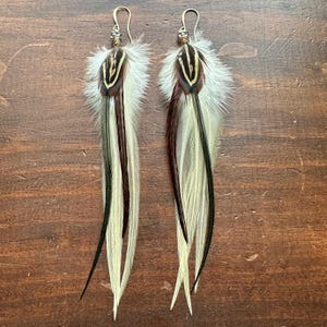 May include: A pair of long feather earrings with brown, black, and white feathers. The earrings have gold-toned metal hoops and beads.