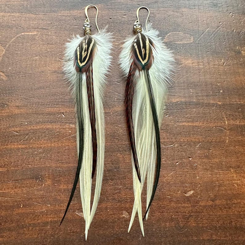 May include: A pair of long feather earrings with brown, black, and white feathers. The earrings have gold-toned metal hoops and beads.