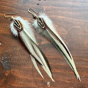May include: A pair of long, white feather earrings with brown and black accents. The earrings are made with natural feathers and have a gold-tone wire.