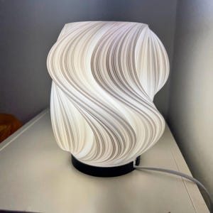 May include: A decorative table lamp with a unique, swirling design. The lamp features a white and gray striped pattern, with a black base. The lamp's shape is organic and modern, and it is lit from within, creating a soft glow.
