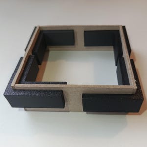 Pode incluir: Estrutura quadrada e aberta com moldura castanha clara e peças de canto pretas. A estrutura parece ser feita de cartão ou material semelhante, com uma superfície texturizada nas peças pretas. O design é simples e geométrico.