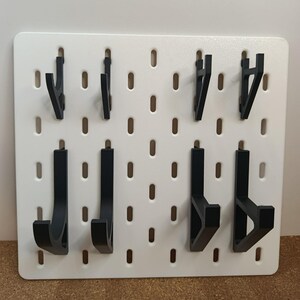 Pode incluir: Um painel perfurado branco com vários ganchos pretos de vários designs. O painel perfurado tem orifícios espaçados uniformemente para colocação de ganchos. Os ganchos são projetados para pendurar itens. Uma ferramenta de organização útil.