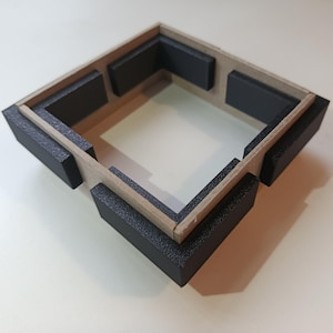 Può includere: Una piccola struttura quadrata aperta, con un telaio in legno marrone chiaro e pezzi angolari neri. L'interno è bianco e la struttura poggia su una superficie di colore chiaro. I pezzi neri hanno un aspetto strutturato.