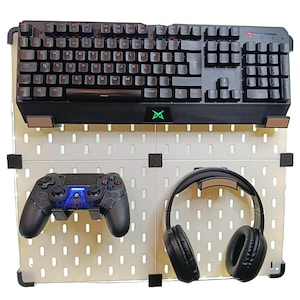 Può includere: Un'attrezzatura da gioco con una tastiera nera, un controller e cuffie su una pannello forato bianco. La tastiera ha tasti illuminati. Il controller ha luci blu. Le cuffie sono di tipo over-ear.