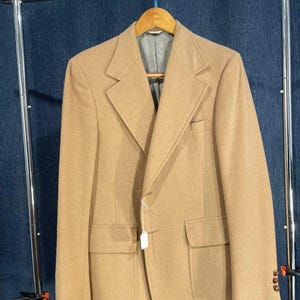 May include: A tan blazer with a single button closure and two flap pockets. The blazer is hanging on a clothes rack.