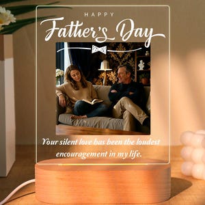 May include: A clear acrylic plaque with a wooden base featuring the text Happy Father's Day and a graphic of a bow tie above a photograph of two people sitting on a sofa. The text Your silent love has been the loudest encouragement in my life is printed below the photo.