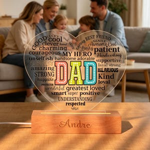 May include: A heart-shaped LED night light with the word "DAD" in colorful letters. The light sits on a wooden base and has the name "Andre" engraved on it. The heart is filled with words like "loving", "kind", and "hero".