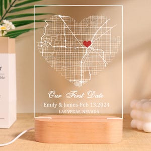 Op de afbeelding: Een heldere acryl nachtlamp met een hartvormige kaart van Las Vegas, Nevada. De tekst "Our First Date" staat boven de namen "Emily & James-Feb 13.2024". Het heeft een houten voet en een wit netsnoer.