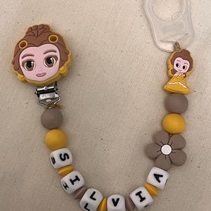 May include: A baby pacifier clip featuring a cartoon princess design. The clip has a metal clasp, a series of beads in yellow, gray, and white, and a flower-shaped bead. The name "Olivia" is spelled out in white letter beads.