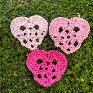 Può includere: Tre applicazioni all'uncinetto a forma di cuore disposte in formazione triangolare. Ogni pezzo presenta un motivo traforato con un bordo smerlato. I cuori sono realizzati in tre distinte tonalità di rosa: rosa pastello chiaro, rosa medio e magenta vivace.