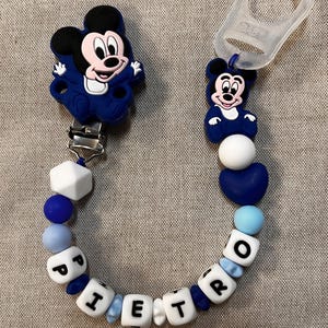 May include: A blue and white pacifier clip with Mickey Mouse motifs. The clip features white lettered beads spelling "PIETRO", interspersed with blue and white beads. Designed to attach to a pacifier, it is a baby accessory.