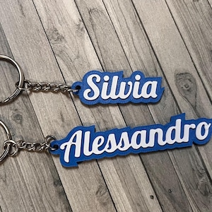 May include: Two custom keychains featuring white script lettering on a blue background. The top keychain displays the name Silvia, and the bottom keychain displays the name Alessandro. Each keychain includes a silver metal ring and a short chain.