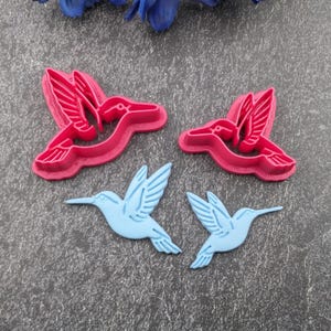 May include: Two pink hummingbird-shaped cookie cutters and two light blue hummingbird-shaped cookie stamps. The cutters feature an open center design with detailed wing and tail impressions, while the stamps are solid silhouettes of hummingbirds.