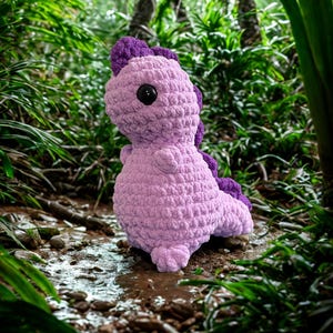 May include: A handmade crochet dinosaur in shades of purple, with a textured body and large black eyes. The dinosaur has a purple crest and is posed in a natural outdoor setting with lush green foliage and a small stream.