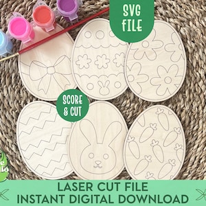 Op de afbeelding: Zes onafgewerkte houten paaseivormige vormen met verschillende ontwerpen, waaronder een konijn, wortels, bloemen en zigzagpatronen. De eieren zijn klaar om te worden beschilderd en versierd. De afbeelding bevat ook verfpotten en een kwast.