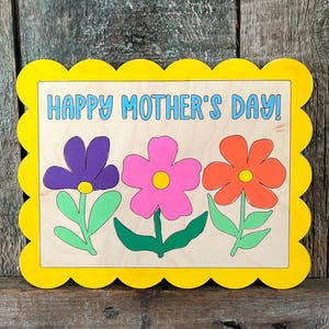 May include: A wooden sign with a scalloped yellow frame and the text "Happy Mother's Day!" in blue. Three flowers are painted on the sign: a purple flower, a pink flower, and an orange flower.