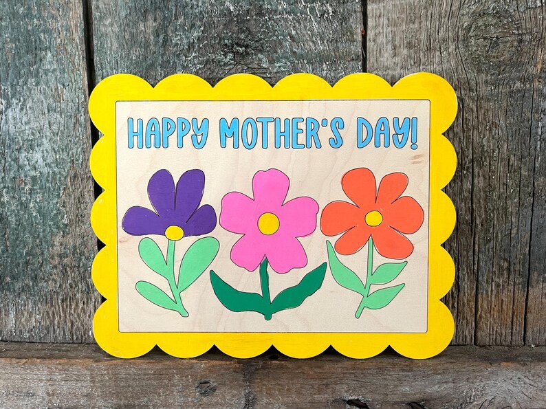 May include: A wooden sign with a scalloped yellow frame and the text "Happy Mother's Day!" in blue. Three flowers are painted on the sign: a purple flower, a pink flower, and an orange flower.