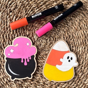 May include: Halloween-themed wooden decorations featuring a black cauldron with pink potion and a candy corn with a ghost. Two black markers with orange and pink tips are also present. The items are arranged on a woven surface.