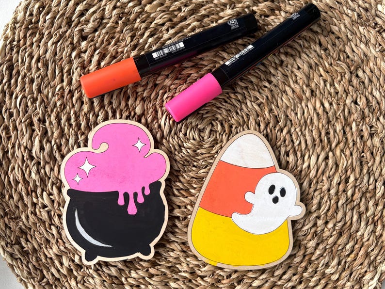 May include: Halloween-themed wooden decorations featuring a black cauldron with pink potion and a candy corn with a ghost. Two black markers with orange and pink tips are also present. The items are arranged on a woven surface.