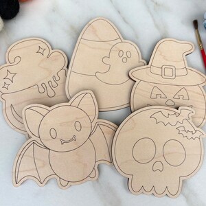 May include: A collection of unfinished wooden Halloween-themed cutouts. The shapes include a cauldron, candy corn with a ghost, a jack-o'-lantern, a bat, and a skull. A red paintbrush and small paint pots are visible in the background.
