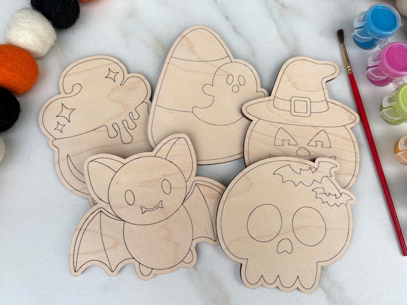 May include: A collection of unfinished wooden Halloween-themed cutouts. The shapes include a cauldron, candy corn with a ghost, a jack-o'-lantern, a bat, and a skull. A red paintbrush and small paint pots are visible in the background.