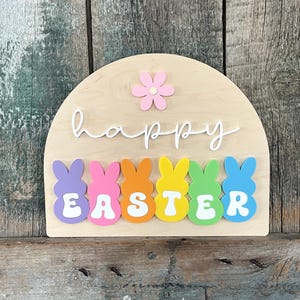 May include: A wooden sign shaped like an arch with the words "happy easter" in white script.  The sign is decorated with colourful pastel bunny shapes and a pink flower.