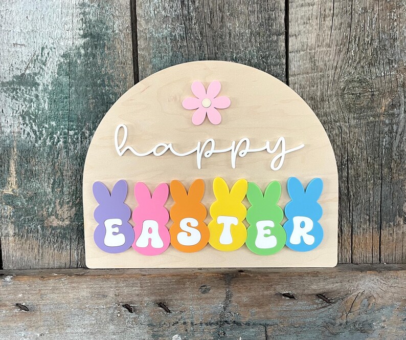 May include: A wooden sign shaped like an arch with the words "happy easter" in white script.  The sign is decorated with colourful pastel bunny shapes and a pink flower.