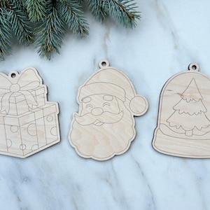 May include: Three unfinished wooden Christmas ornaments. One is a stack of presents with a bow, another is Santa Claus, and the third is a snow globe with a Christmas tree inside. Each ornament has a hole for hanging.