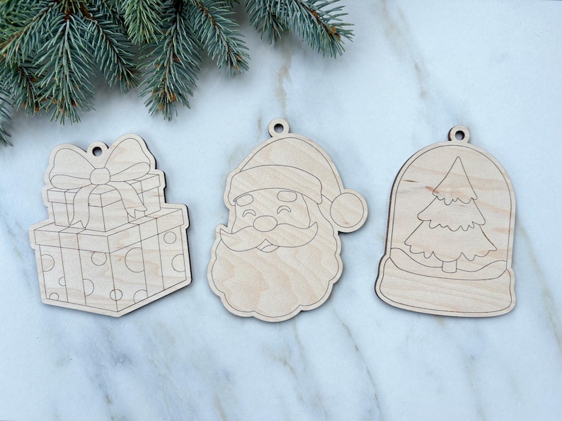 May include: Three unfinished wooden Christmas ornaments. One is a stack of presents with a bow, another is Santa Claus, and the third is a snow globe with a Christmas tree inside. Each ornament has a hole for hanging.