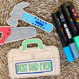 May include: Wooden cutouts of a hammer, saw, and toolbox with the text "BEST DAD EVER". The hammer has a gray head and blue handle. The saw is red and gray. The toolbox is light brown. Also, there are five colorful markers.