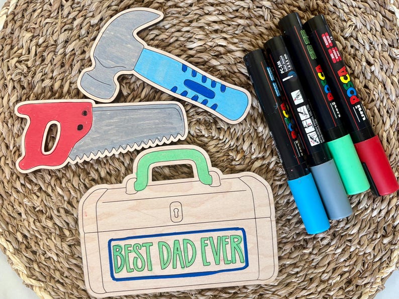 May include: Wooden cutouts of a hammer, saw, and toolbox with the text "BEST DAD EVER". The hammer has a gray head and blue handle. The saw is red and gray. The toolbox is light brown. Also, there are five colorful markers.