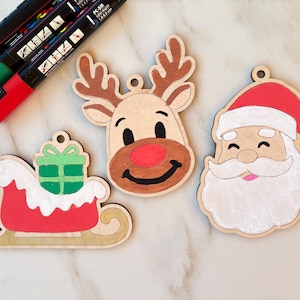 May include: Wooden Christmas ornaments featuring a reindeer, Santa Claus, and a sleigh with a gift. The ornaments are hand-painted with red, brown, green, and white accents. Paint pens are visible in the upper left corner. Each ornament has a small hole for hanging.