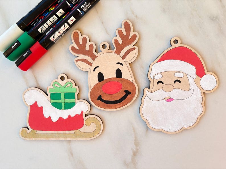 May include: Wooden Christmas ornaments featuring a reindeer, Santa Claus, and a sleigh with a gift. The ornaments are hand-painted with red, brown, green, and white accents. Paint pens are visible in the upper left corner. Each ornament has a small hole for hanging.