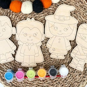 May include: Four unfinished wooden cutouts featuring cartoon characters including a monster, a vampire, a witch, and a mummy. Below the wooden shapes is a strip of six small plastic paint pots containing blue, pink, green, yellow, black, and white paint.
