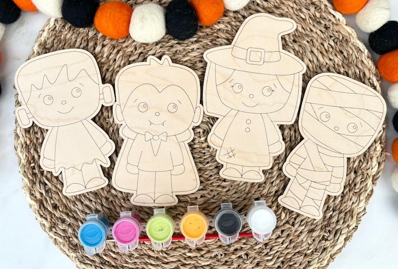 May include: Four unfinished wooden cutouts featuring cartoon characters including a monster, a vampire, a witch, and a mummy. Below the wooden shapes is a strip of six small plastic paint pots containing blue, pink, green, yellow, black, and white paint.