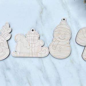 May include: Four wooden Christmas ornaments: a reindeer head, a sleigh with a gift, a snowman, and a candy cane. Each ornament has a small hole for hanging. The ornaments are made of light-colored wood.