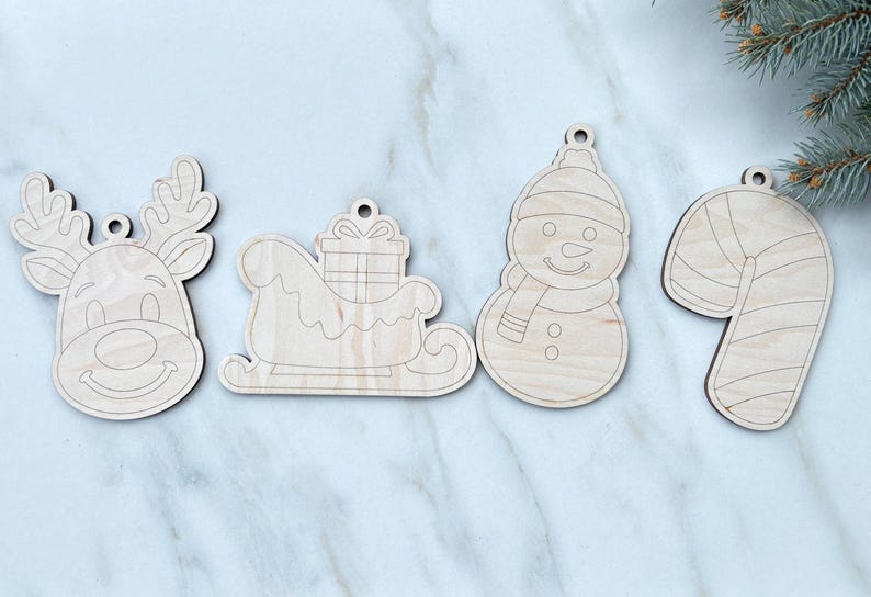 May include: Four wooden Christmas ornaments: a reindeer head, a sleigh with a gift, a snowman, and a candy cane. Each ornament has a small hole for hanging. The ornaments are made of light-colored wood.