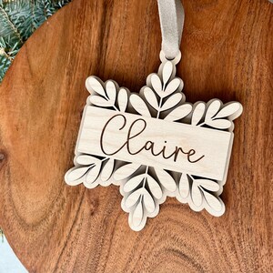 May include: A wooden snowflake ornament with the name "Claire" engraved on it. The ornament is white and has a brown string for hanging.