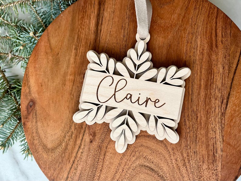 May include: A wooden snowflake ornament with the name "Claire" engraved on it. The ornament is white and has a brown string for hanging.