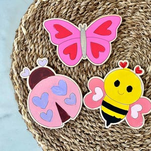 Puede incluir: Tres recortes de madera pintados en rosa, amarillo y morado. Los recortes tienen forma de mariposa, mariquita y abeja. Cada recorte tiene detalles en forma de coraz&oacute;n.