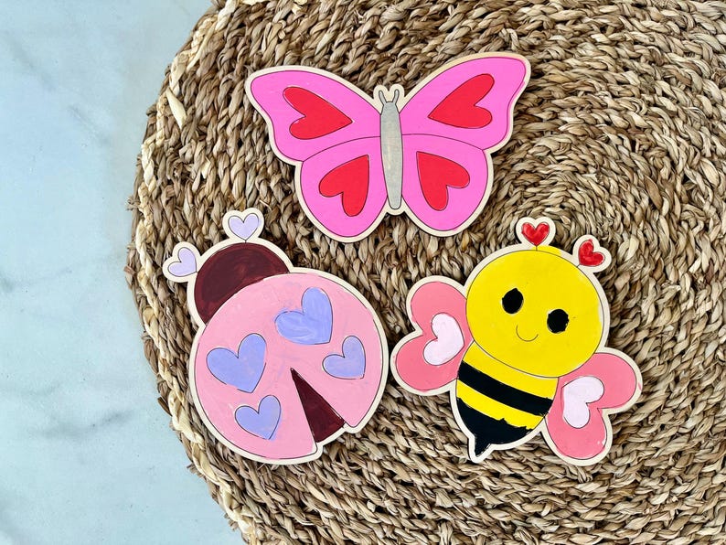 Puede incluir: Tres recortes de madera pintados en rosa, amarillo y morado. Los recortes tienen forma de mariposa, mariquita y abeja. Cada recorte tiene detalles en forma de coraz&oacute;n.